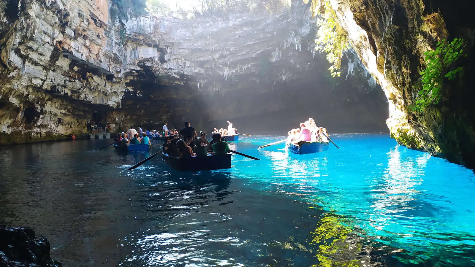 Ruderboote auf dem türkisfarbenen See der Melissani-Höhle auf Kefalonia, Sonnenstrahlen fallen durch das eingestürzte Höhlendach
